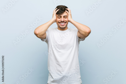 Young caucasian man wearing a white tshirt laughs joyfully keeping hands on head. Happiness concept.