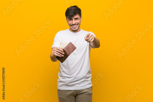 Young hispanic man holding a wallet cheerful smiles pointing to front.