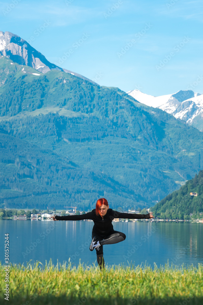 An attractive young woman doing a yoga pose for balance and stretching near the lake Zell am See high in the Alpine mountains in Austria. dressed in a black tracksuit for training, vertical photo