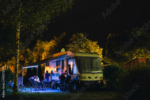 Fototapeta People sit under an awning and watch TV, Night view of the parking lot for a motorhome, camper van, campsite camp for sleeping and relaxing