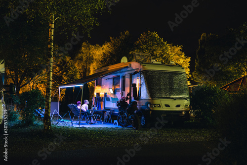 People sit under an awning and watch TV, Night view of the parking lot for a motorhome, camper van, campsite camp for sleeping and relaxing. vacation and travel tour. motorcycle.