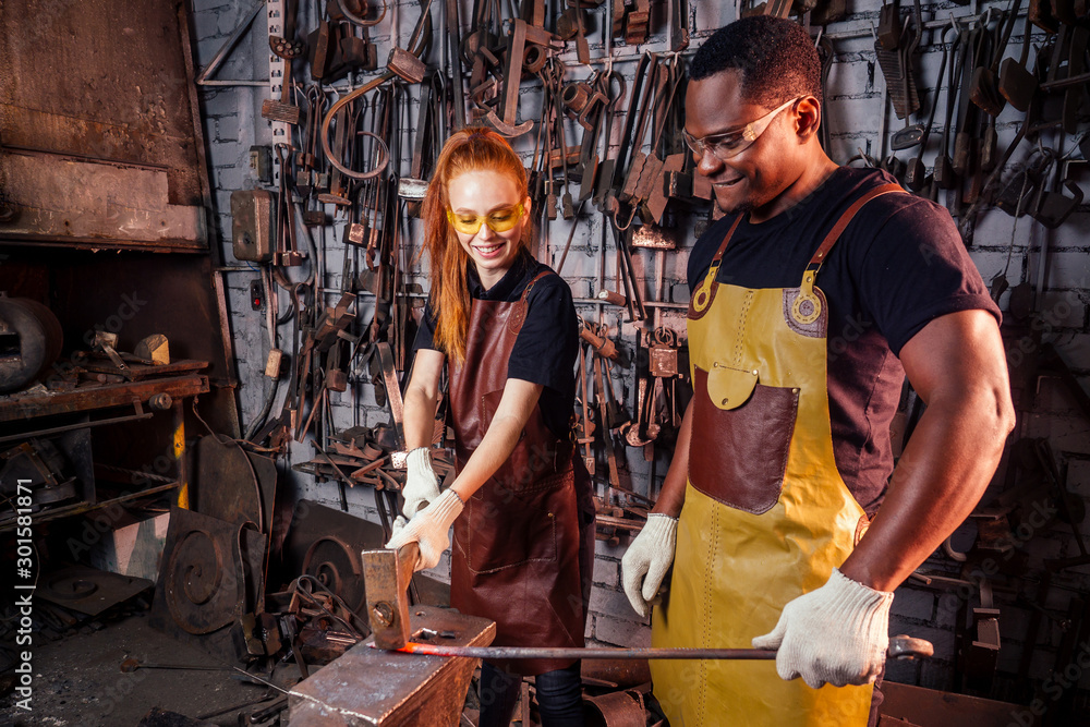 couple in love redhaired ginger young european woman and afro-american man wearing leather apron working blacksmith workshop.small family international business concept