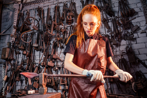 redhaired ginger young european feminist woman wearing leather apron working blacksmith workshop.small business strong and independent concept