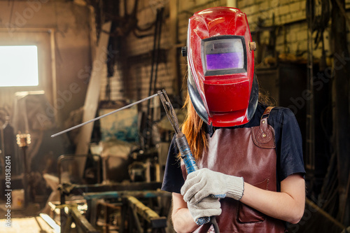 Strong and worthy pretty redhead ginher woman wearing protection helmet and leather apron with gloves holding welding machine instrument . small business concept