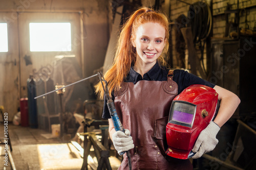 Strong and worthy pretty redhead ginher woman wearing protection helmet and leather apron with gloves holding welding machine instrument . small business concept