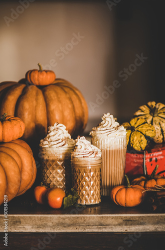 Pumpkin spice latte coffee drink topped with whipped cream and cinnamon in tall glasses among fresh pumpkins and persimmons over kitchen counter, copy space. Seasonal Autumn hot warming sweet drink