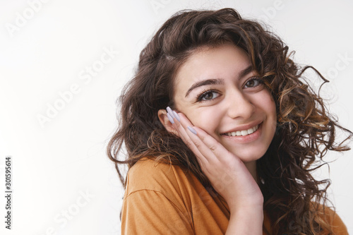 Tender delighted young flirty caucasian girl curly black hair pressing palm cheek blushing nice heartwarming compliment smiling broadly feel upbeat in love, standing passionately gazing camera