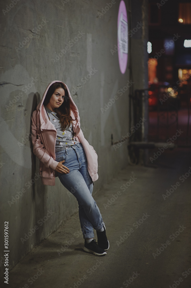 Naklejka premium A beautiful girl in a pink sweater with a hood stands against the backdrop of the night city with a smartphone in her hand. Soft focus, colored highlights in the background.