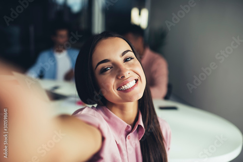 Close up portrait of cheerful hispter girl with perfect smile looking at camera and taking selfie photos during leisure time indoors, happy female teenager with brunette hair enjoying joyful mood