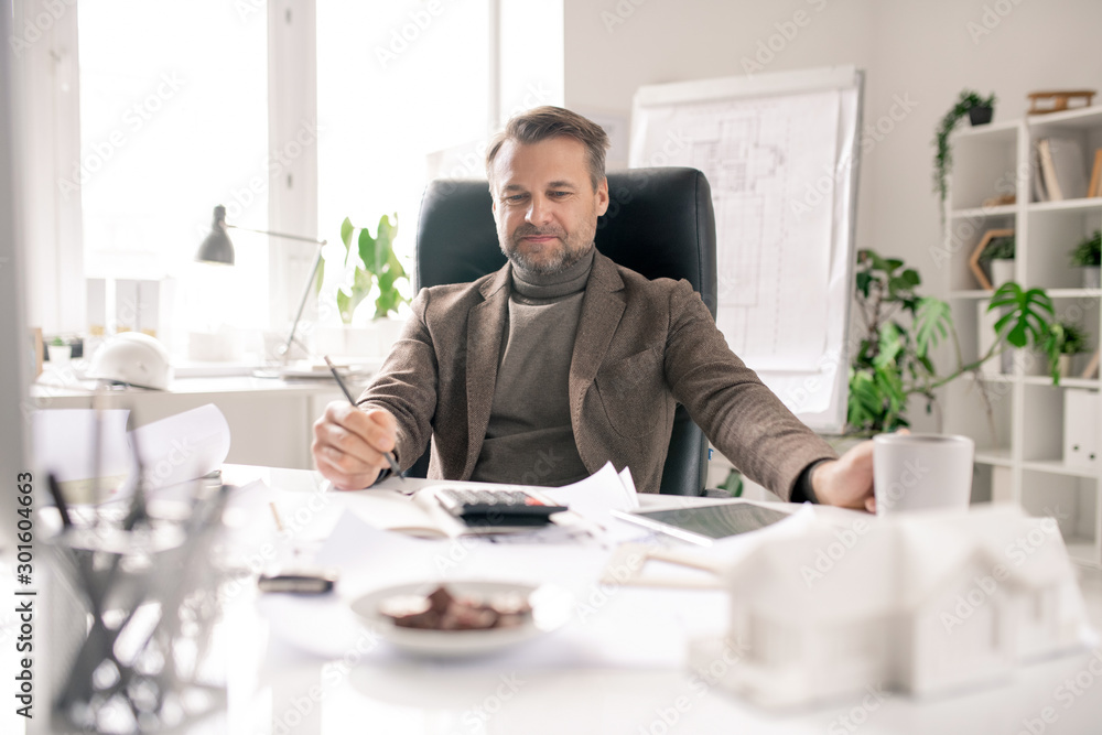 Mature accountant with pen and cup of coffee making calculations by workplace