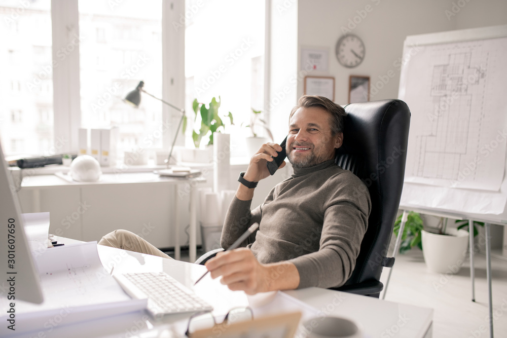 Cheerful mature businessman with smartphone looking at you with smile by desk Cheerful mature businessman with smartphone looking at you with smile by desk