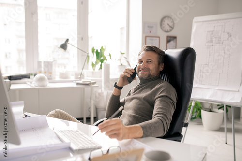 Cheerful mature businessman with smartphone looking at you with smile by desk