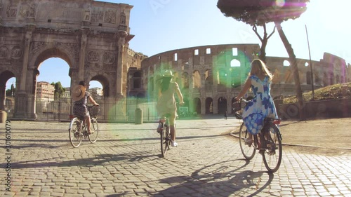Three happy young women friends tourists riding bikes at Colosseum in Rome, Italy at sunrise.