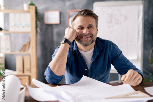 Smiling mature engineer sitting by table while working over new sketches
