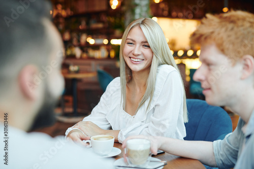 Happy blonde girl with toothy smile having cup of cappuccino with two guys