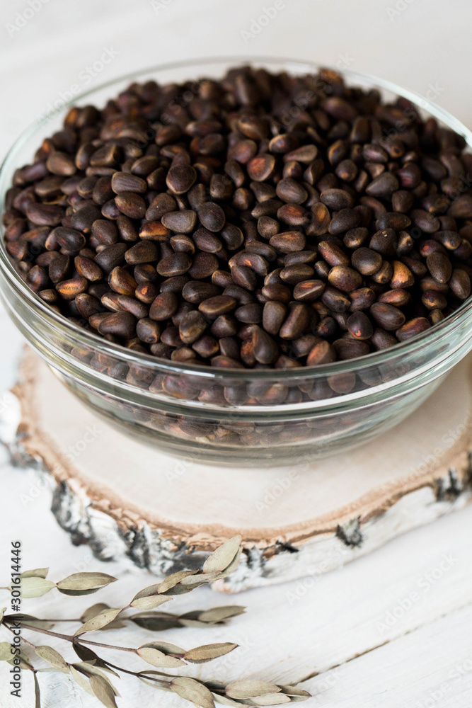 On a light wooden table on a birch saw-board there is a glass transparent deep plate full of fresh pine nuts in the shell. On a light wooden table on a birch saw-board there is a glass transparent deep plate full of fresh pine nuts in the shell.