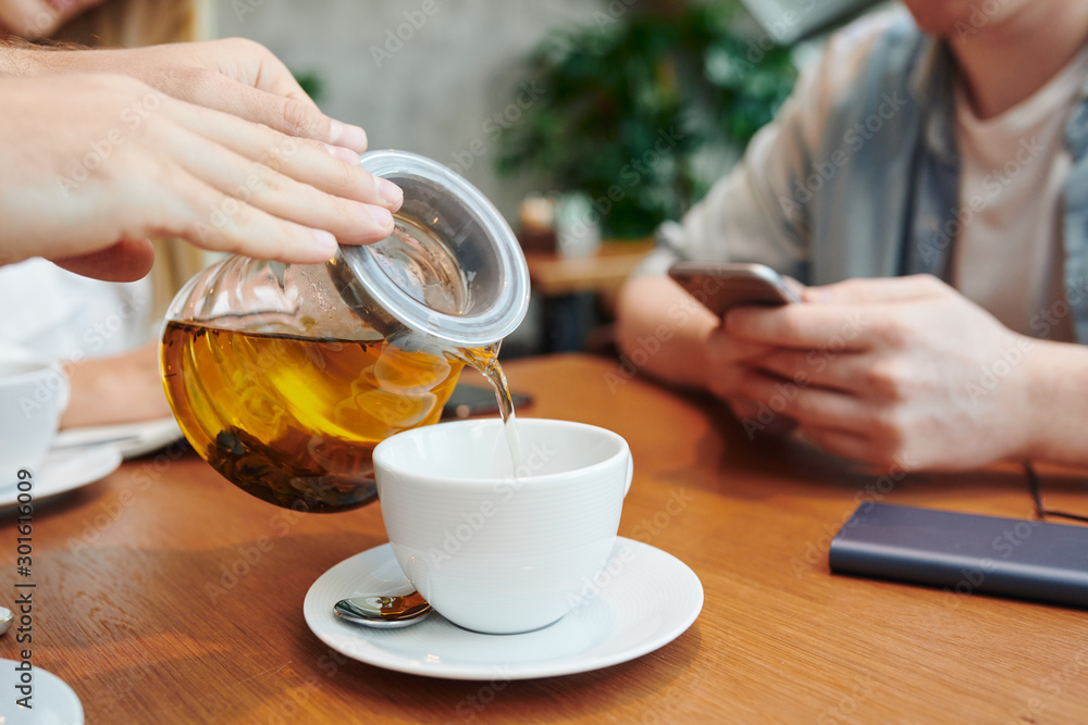 Hands of guy with teapot pouring herbal tea into white porcelain cup in cafe Hands of guy with teapot pouring herbal tea into white porcelain cup in cafe