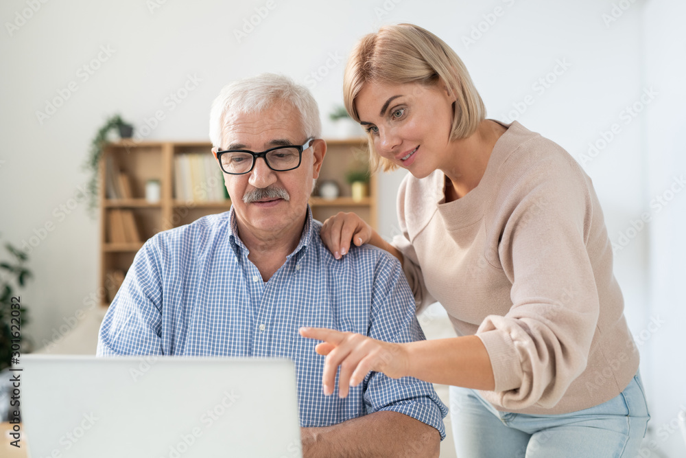 Young woman pointing at laptop display while explaining something to her father - obrazy, fototapety, plakaty 