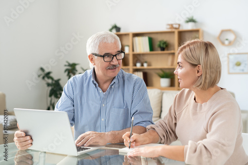 Young woman making notes in notepad while looking at senior father with laptop