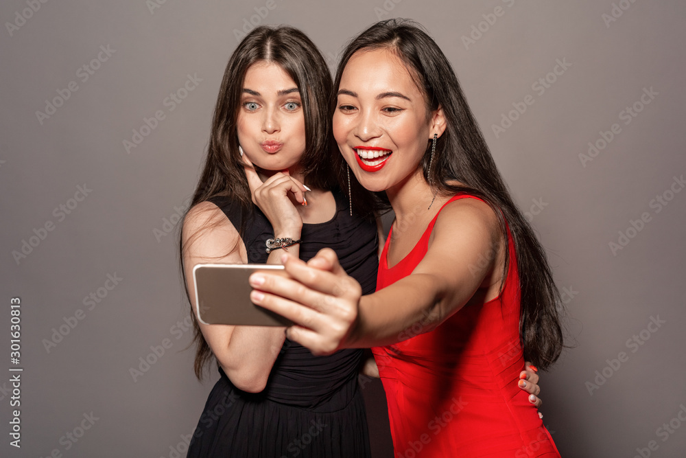Freestyle. Young girls in dress standing hugging isolated on gray taking selfie on smartphone grimacing cute Freestyle. Young girls in dress standing hugging isolated on gray taking selfie on smartphone grimacing cute