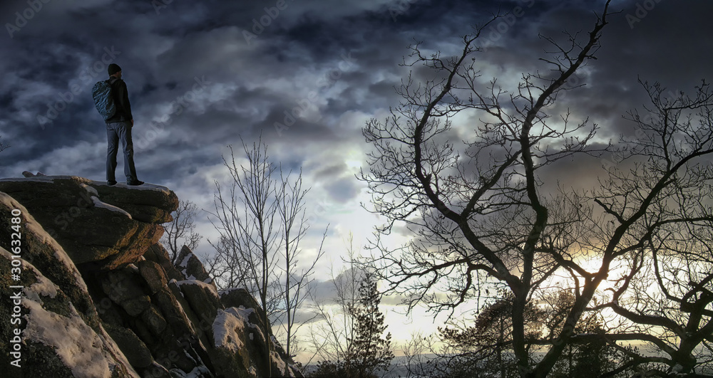 Male hiker with backpack on a mountain range cliff behind a city looking at rugged winter landscape enjoying weekend.Old trees, relaxing, freedom, silence and calm,sunset,loneliness.Composite photo. .