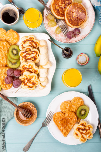 Healthy breakfast with fresh hot waffles hearts, pancakes flowers with berry jam and fruits on turquoise background, top view, flat lay. Food concept.