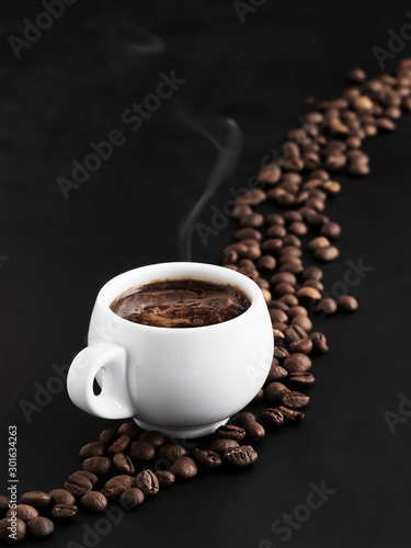 White cup with fragrant espresso on a black background, steam rises above the cup. Roasted coffee beans are located around a cup of coffee and in the background. Close-up. © Ed