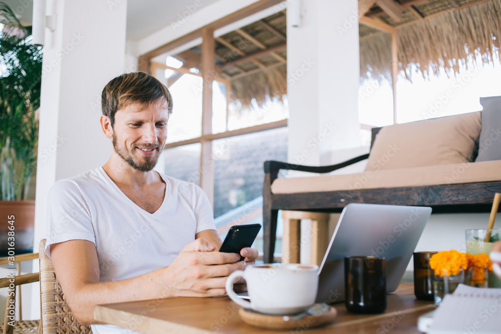 Smiling bearded man texting on cellphone during break in home office Smiling bearded man texting on cellphone during break in home office