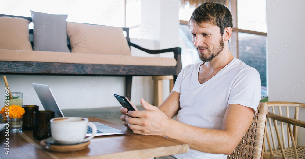 Positive hipster guy reading funny notification on smartphone sitting front laptop computer in cafe, successful male sending money via online banking app on cellphone connected to public wifi internet