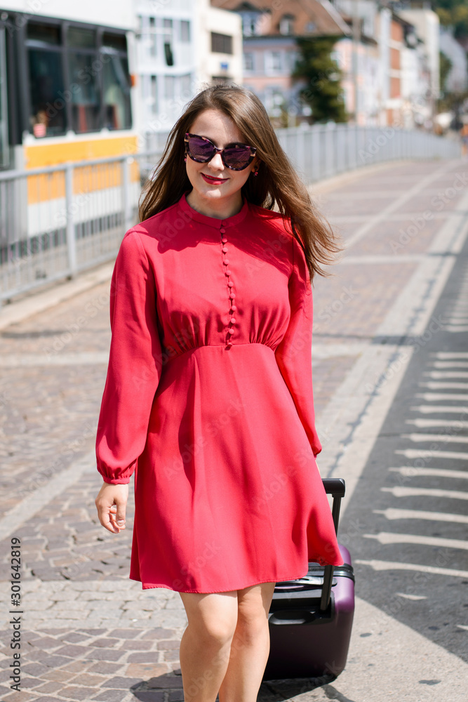 Young girl dressed in fashionable red dress carrying luggage. Female traveler arrived to some other city for weekends to have shopping and spend time with friends. Cheerful girl likes travelling