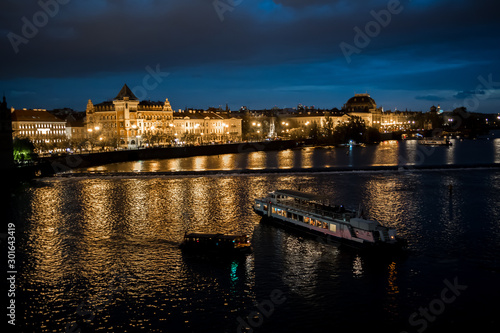 Illuminated Moldova River With Ship And Historic Buildings In The Night In Prague In The Czech Republic