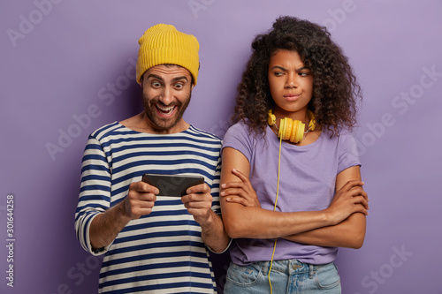 Addicted hipster guy plays smartphone, ignores girlfriend, Afro female gets bored, keeps arms crossed and looks negatively. Man in yellow hat and striped jumper obsessed with modern technologies