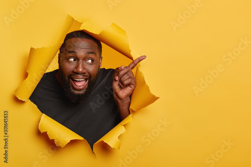 Photo of happy cheerful black male model points index finger aside, keeps mouth opened, shows tongue, attracts your attention to blank space, invites you going there, recommends product. Advertisement © wayhome.studio 