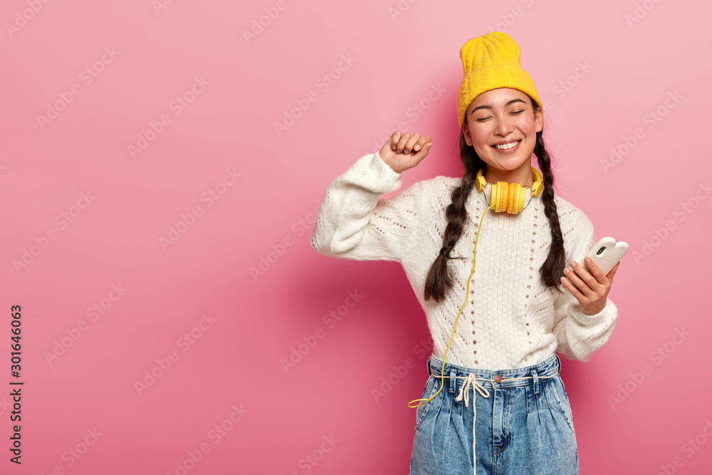 Tender modern woman in stylish clothing enjoys awesome beat in headphones, dances with rhythm of music with hand raised, holds smartphone in hand, smiles joyfully, isolated over pink background Tender modern woman in stylish clothing enjoys awesome beat in headphones, dances with rhythm of music with hand raised, holds smartphone in hand, smiles joyfully, isolated over pink background