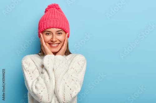 Romantic positive young European woman smiles gently, has white perfect teeth, touches both cheeks, has friendly look, wears pink hat with pompon and white sweater, models against blue wall.