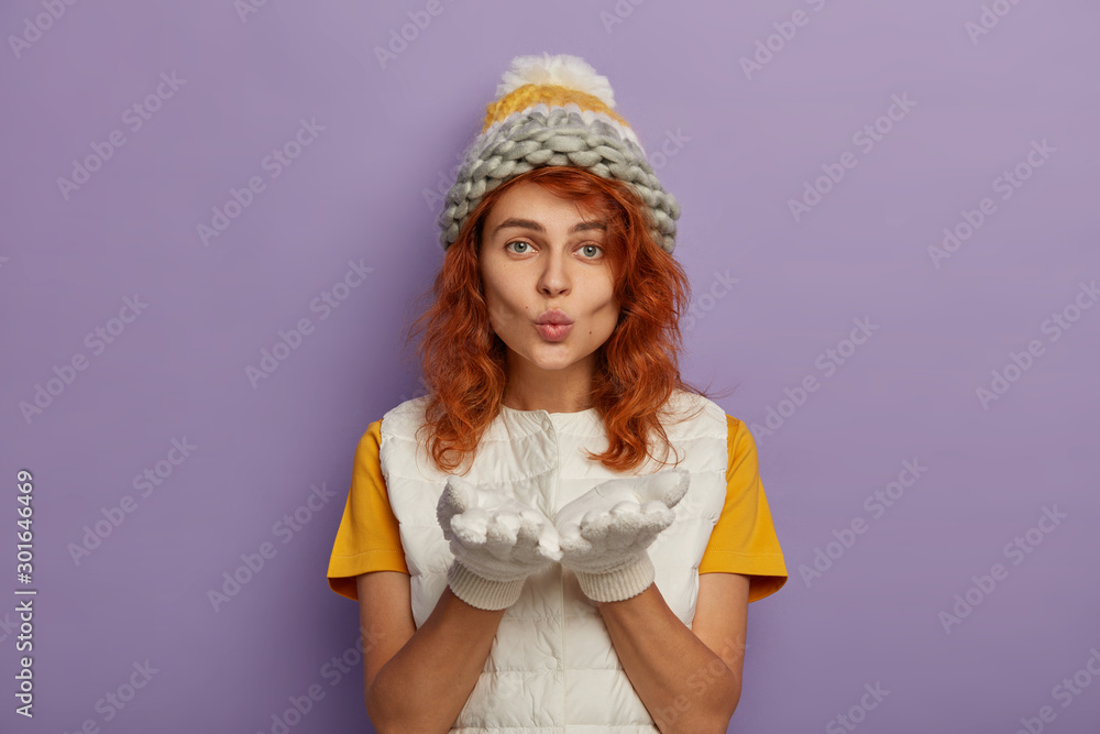 Φωτογραφία Redhead woman blows mwah at camera, keeps palms forward, wears knitted hat and white vest, enjoys winter time, sends air kiss, isolated over purple background
