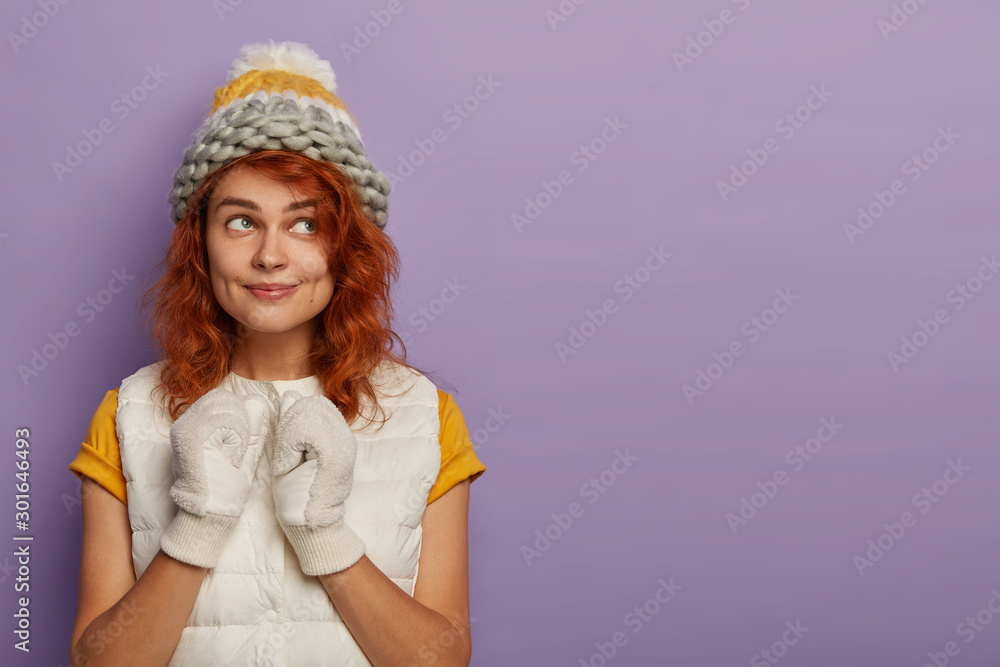 Φωτογραφία Content lovely Caucasian foxy woman wears winter white hat, gloves, dreams about nice vacation during holidays, keeps gaze aside, poses against purple background, copy space for your promotion