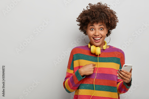 Horizontal shot of pretty woman points at herself, happy being chosen for promotion, holds modern cellphone, yellow headphones on neck, wears striped sweater, isolated on white wall, blank space