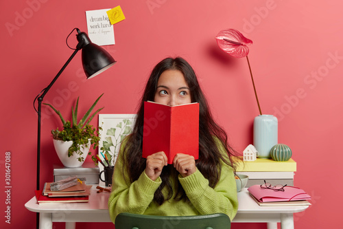 Beautiful brunette pensive mixed race female student learns information from textbook, covers half of face with red diary, poses in coworking space against desktop with necessary things for studying photo