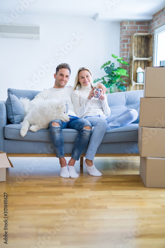 Young beautiful couple with dog sitting on the sofa drinking coffee at new home around cardboard boxes