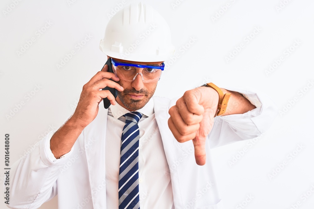 Young scientist man wearing safety helmet and calling using smartphone over isolated background with angry face, negative sign showing dislike with thumbs down, rejection concept Young scientist man wearing safety helmet and calling using smartphone over isolated background with angry face, negative sign showing dislike with thumbs down, rejection concept