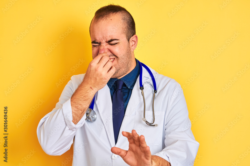 Young doctor man wearing coat and stethoscope standing over isolated yellow background smelling something stinky and disgusting, intolerable smell, holding breath with fingers on nose. Bad smell Young doctor man wearing coat and stethoscope standing over isolated yellow background smelling something stinky and disgusting, intolerable smell, holding breath with fingers on nose. Bad smell