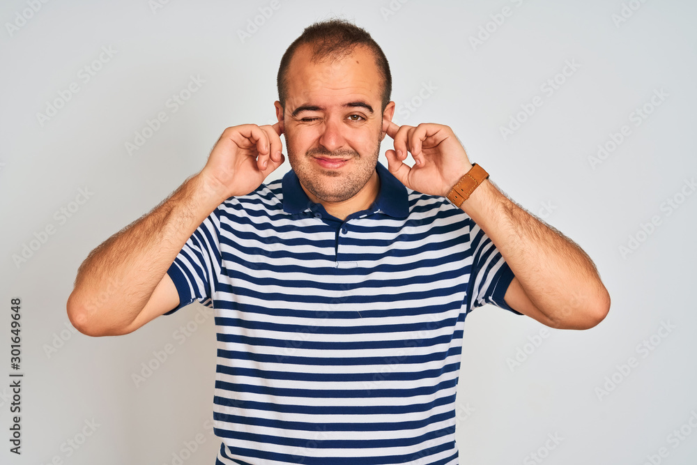 Young man wearing casual striped polo standing over isolated white background covering ears with fingers with annoyed expression for the noise of loud music. Deaf concept. Young man wearing casual striped polo standing over isolated white background covering ears with fingers with annoyed expression for the noise of loud music. Deaf concept.