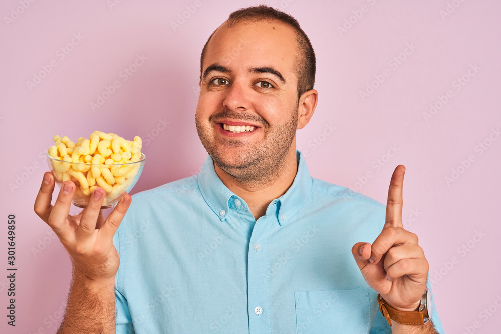 Young man holding bowl with extruded corn standing over isolated pink background surprised with an idea or question pointing finger with happy face, number one Young man holding bowl with extruded corn standing over isolated pink background surprised with an idea or question pointing finger with happy face, number one