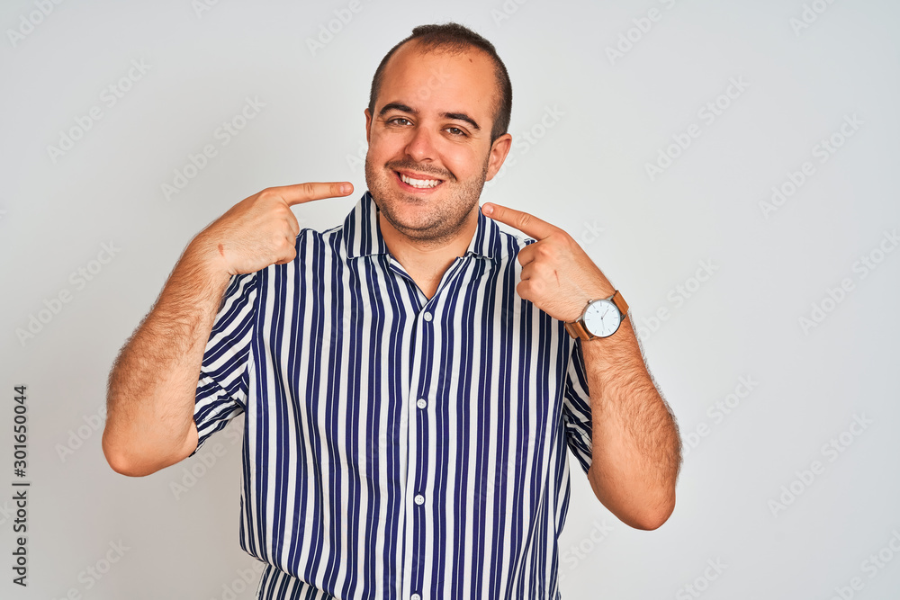 Young man wearing blue striped shirt standing over isolated white background smiling cheerful showing and pointing with fingers teeth and mouth. Dental health concept. Young man wearing blue striped shirt standing over isolated white background smiling cheerful showing and pointing with fingers teeth and mouth. Dental health concept.