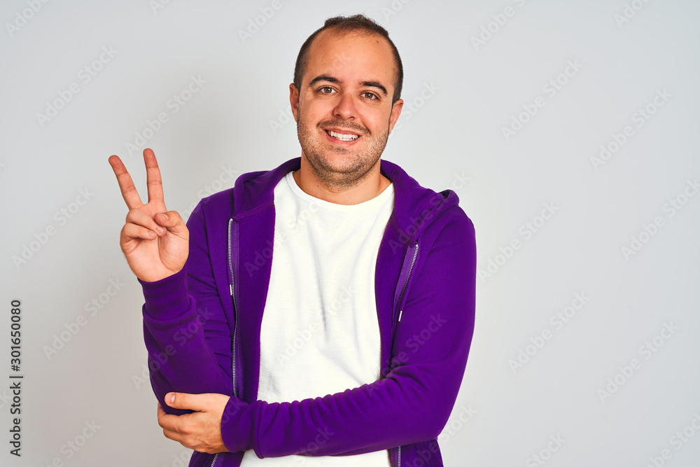 Young man wearing purple sweatshirt standing over isolated white background smiling with happy face winking at the camera doing victory sign. Number two. Young man wearing purple sweatshirt standing over isolated white background smiling with happy face winking at the camera doing victory sign. Number two.