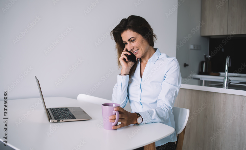 Woman talking on phone holding coffee working on laptop at home Woman talking on phone holding coffee working on laptop at home