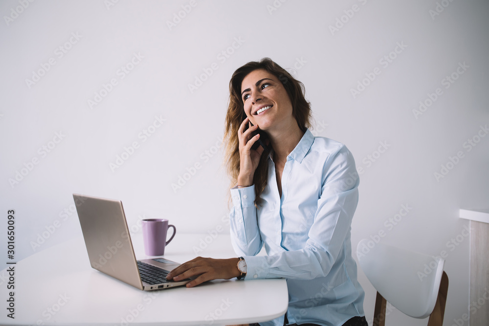 Smiling female talking on mobile phone sitting with computer and coffee at table Smiling female talking on mobile phone sitting with computer and coffee at table
