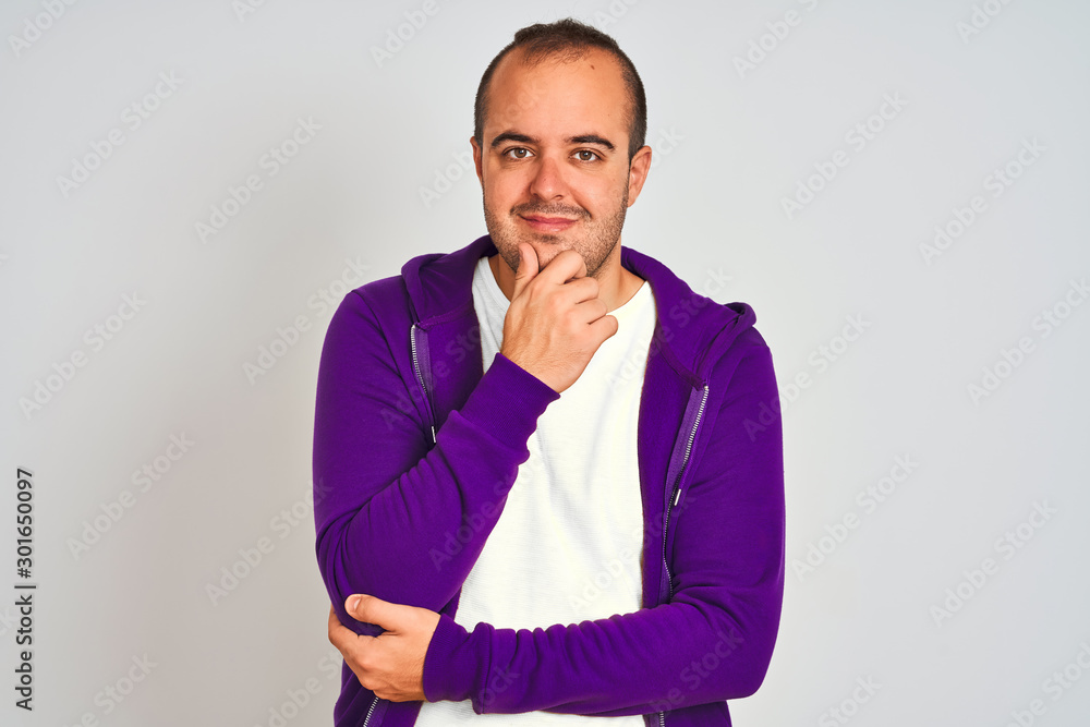 Young man wearing purple sweatshirt standing over isolated white background looking confident at the camera smiling with crossed arms and hand raised on chin. Thinking positive. Young man wearing purple sweatshirt standing over isolated white background looking confident at the camera smiling with crossed arms and hand raised on chin. Thinking positive.