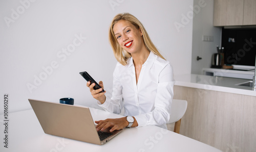 Smiling young blonde with smartphone and laptop looking at camera